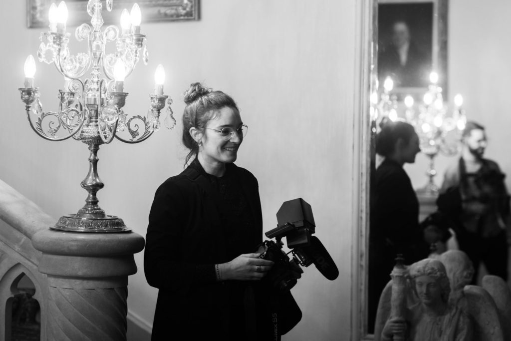 A videographer, camera in hand, smiles with her colleague in the mirror